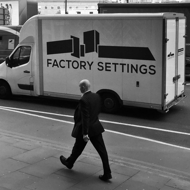 A black and white photo of a London businessman striding robotically in front of a truck with the words ‘FACTORY SETTINGS’ on the side