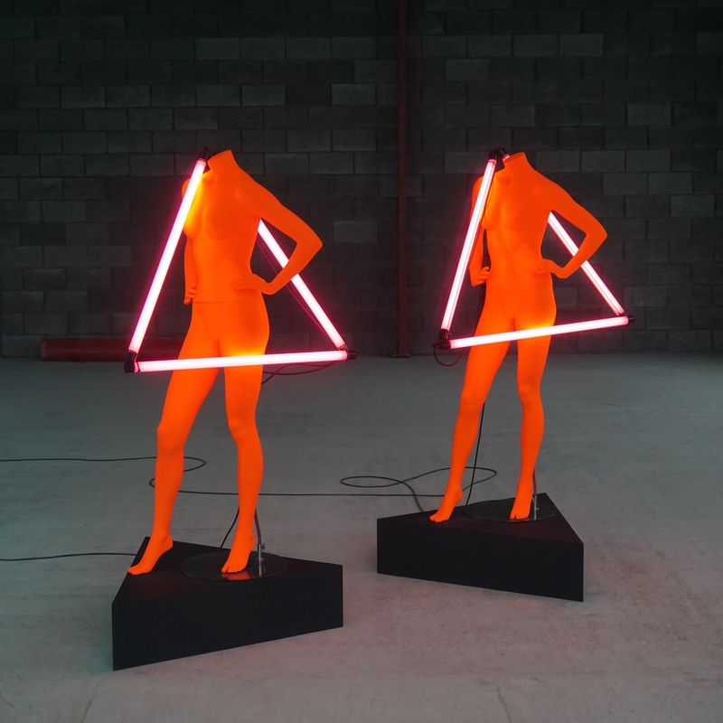 A photo of two naked, headless, red shop dummys with neon triangles hung over their shoulders