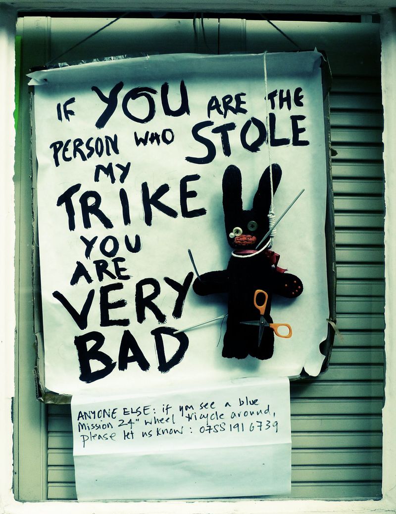 A photo of an angry sign in a window in Brighton. The sign reads ‘If YOU are the person who stole my trike you are VERY bad’. Hanging by its neck in front of the sign is a home made voodoo doll with scissors sticking in it