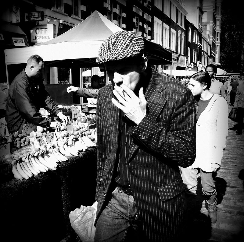 A black and white photo of a guy in a flat cap and pinstripe suit, smoking a cigarette, walking through Strutton Ground market in London