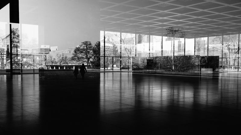 A black and white photo of the interior of an empty office lobby. The reflections on the windows have created overlayed images